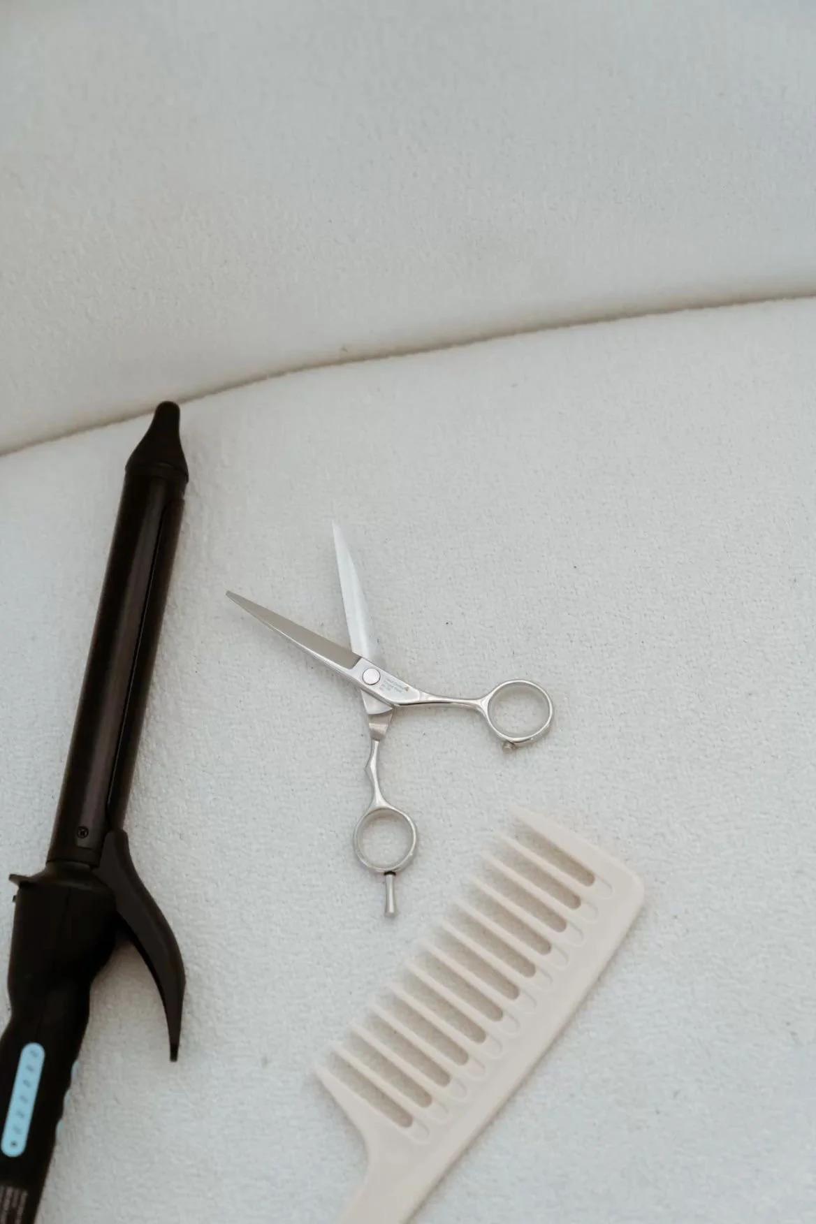 Haircutting tools including a black curling iron, a pair of silver scissors, and a white hair comb resting on a white surface.