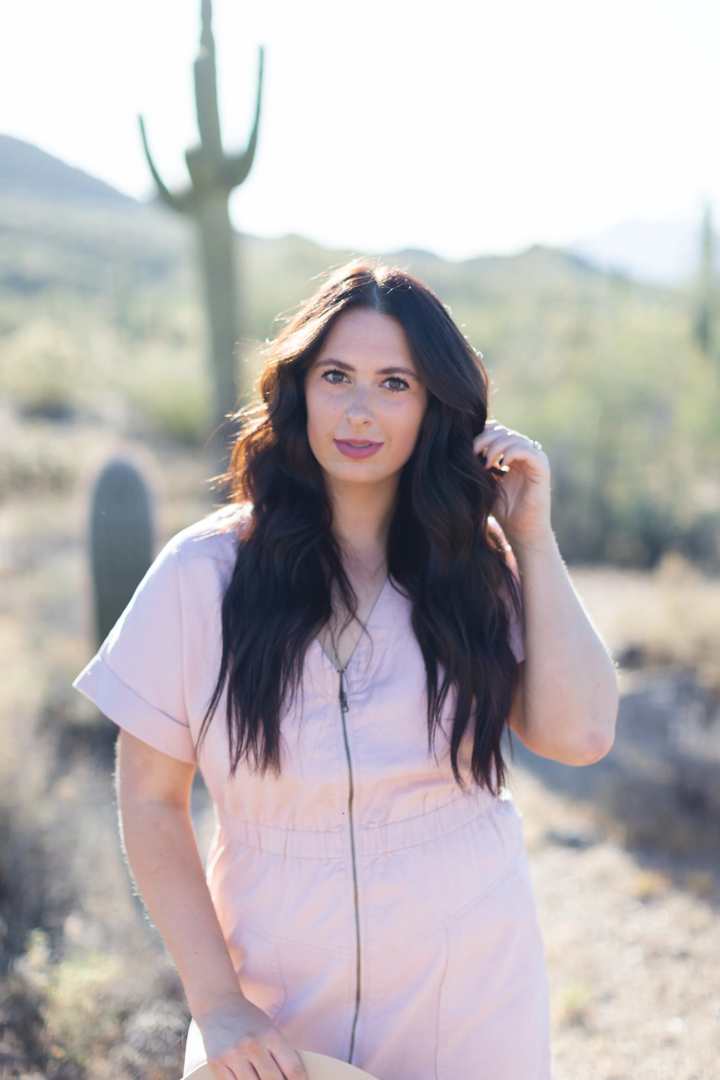 A woman with long dark hair standing outdoors in a sunny desert landscape with cacti and mountains in the background.