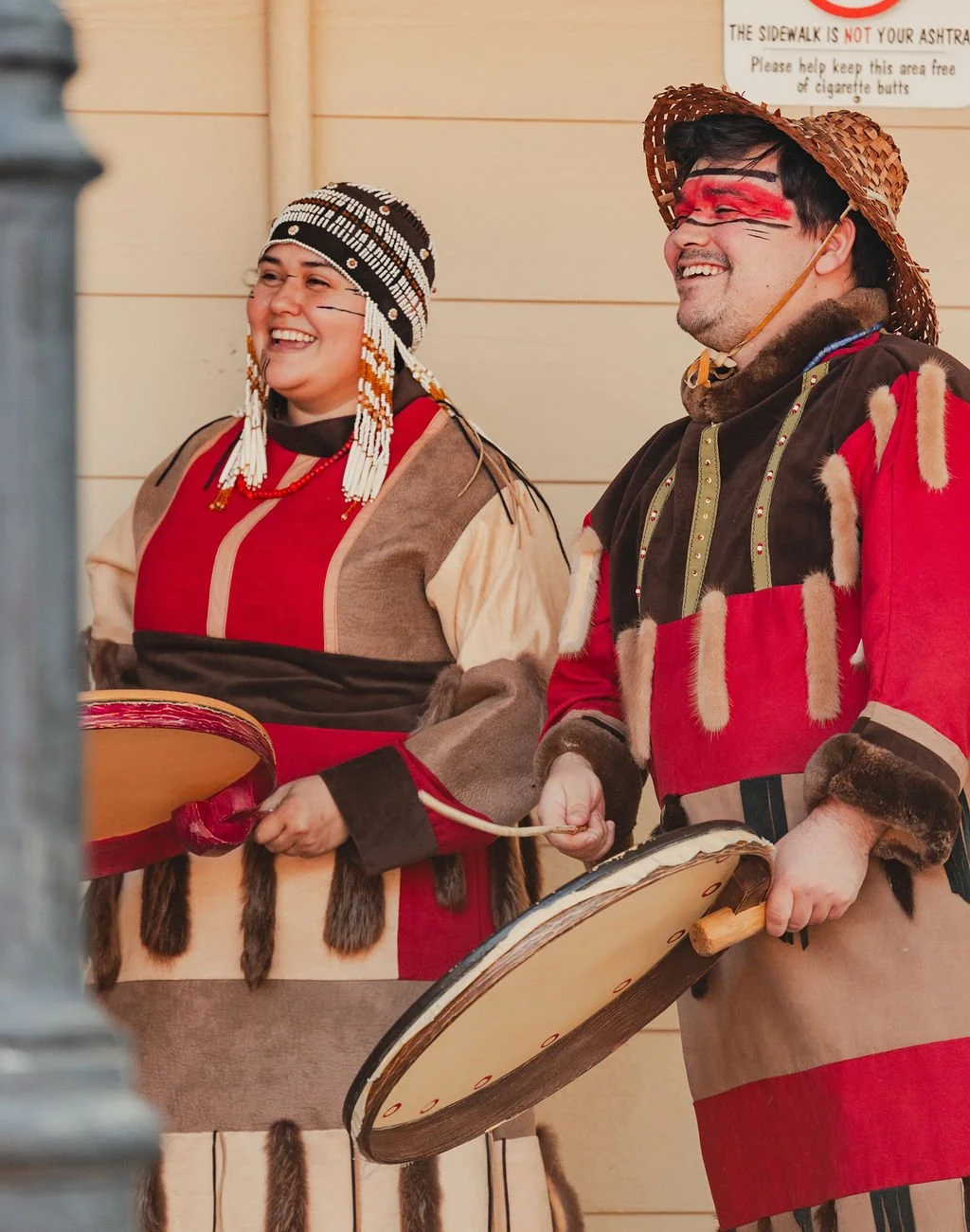 A couple of dancers are photographed smiling during a performance at Kodiak Crab Festival. Both dancers play traditional skin drums and wear furs and face paint.