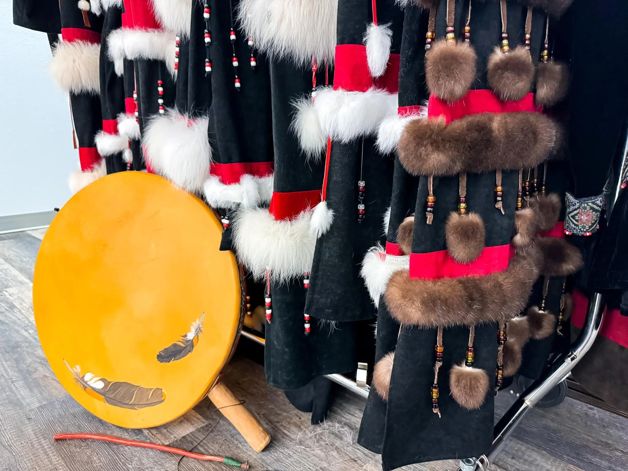 Hanging Kodiak Alutiiq Dancers regalia with a yellow drum in front. Regalia is traditional clothing worn during dances and feature feathers, skins, and beads.