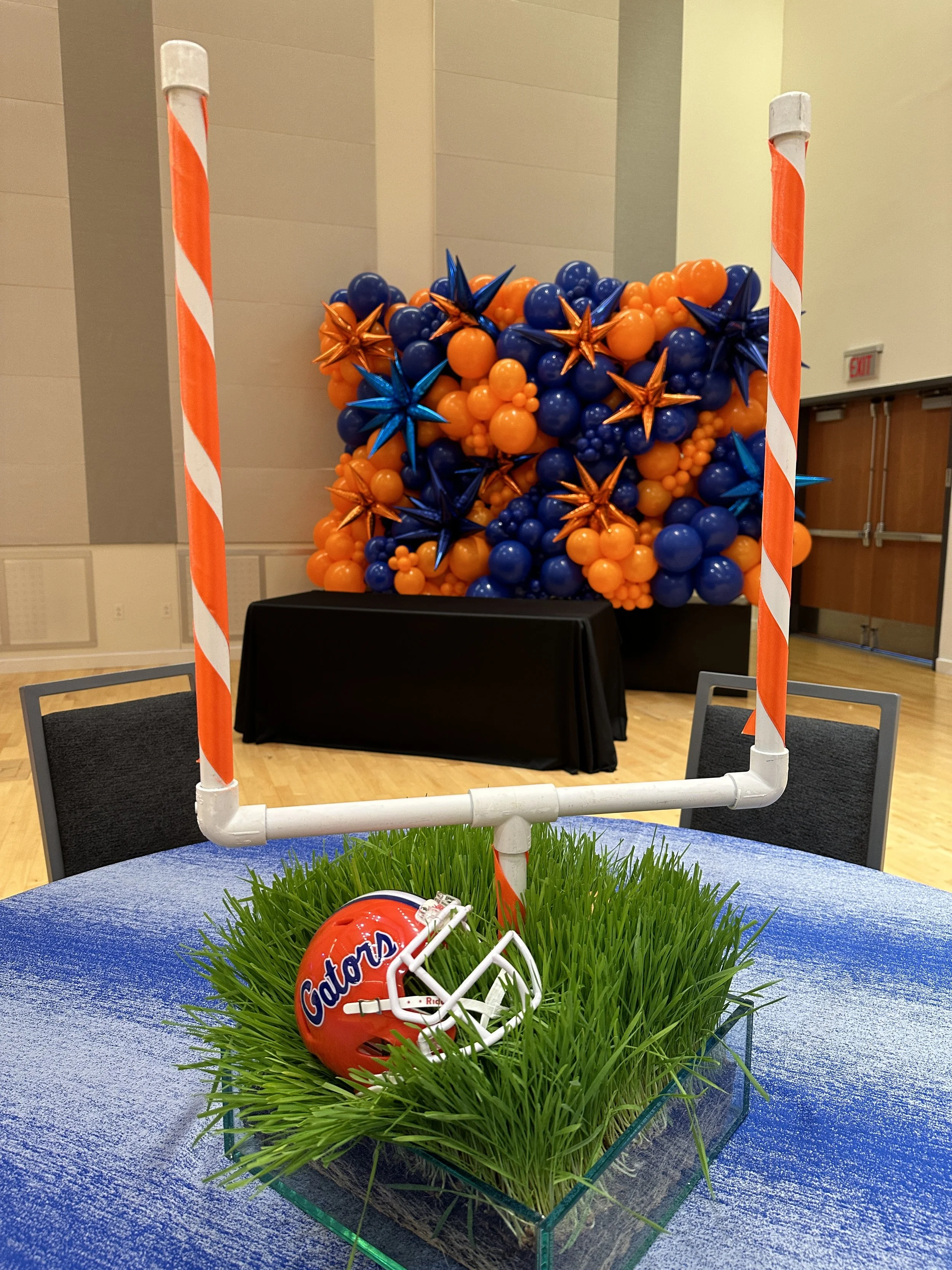 Football helmet with 'Gators' logo placed among green grass on a table, with a backdrop of orange and blue balloons arranged as a decorative display behind a barricade.