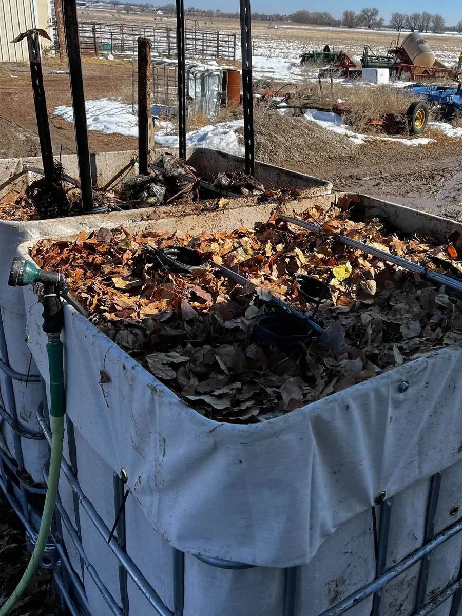 White IBC tote filled with brown autumn leaves on a snowy farmyard, tractors in the background