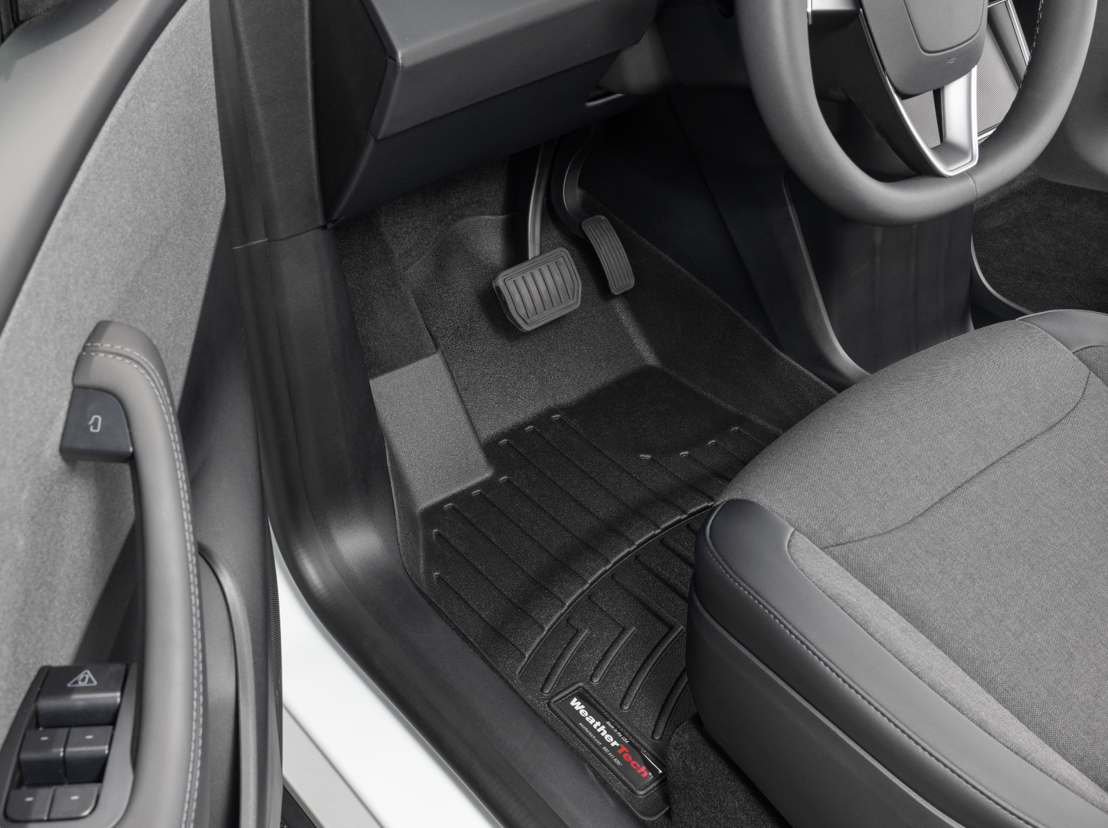 The image shows the driver side interior of a vehicle, focusing on the pedals, footwell, and a floor mat with the WeatherTech logo. The pedals are for acceleration and braking, and part of the steering wheel and seat are also visible.