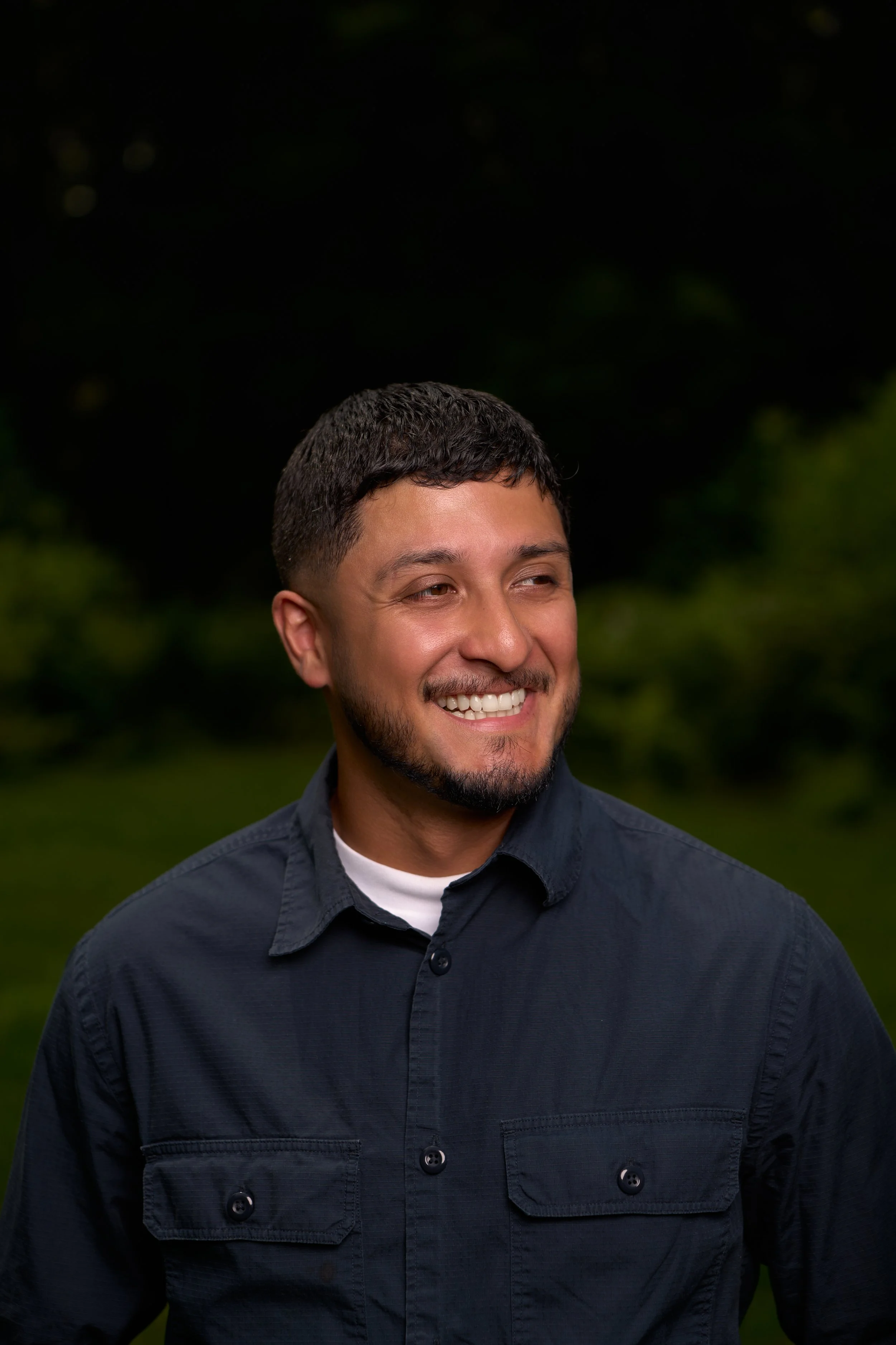 A young man with short, dark hair and a beard, smiling and wearing a dark button-up shirt, standing outdoors with a blurred green background.