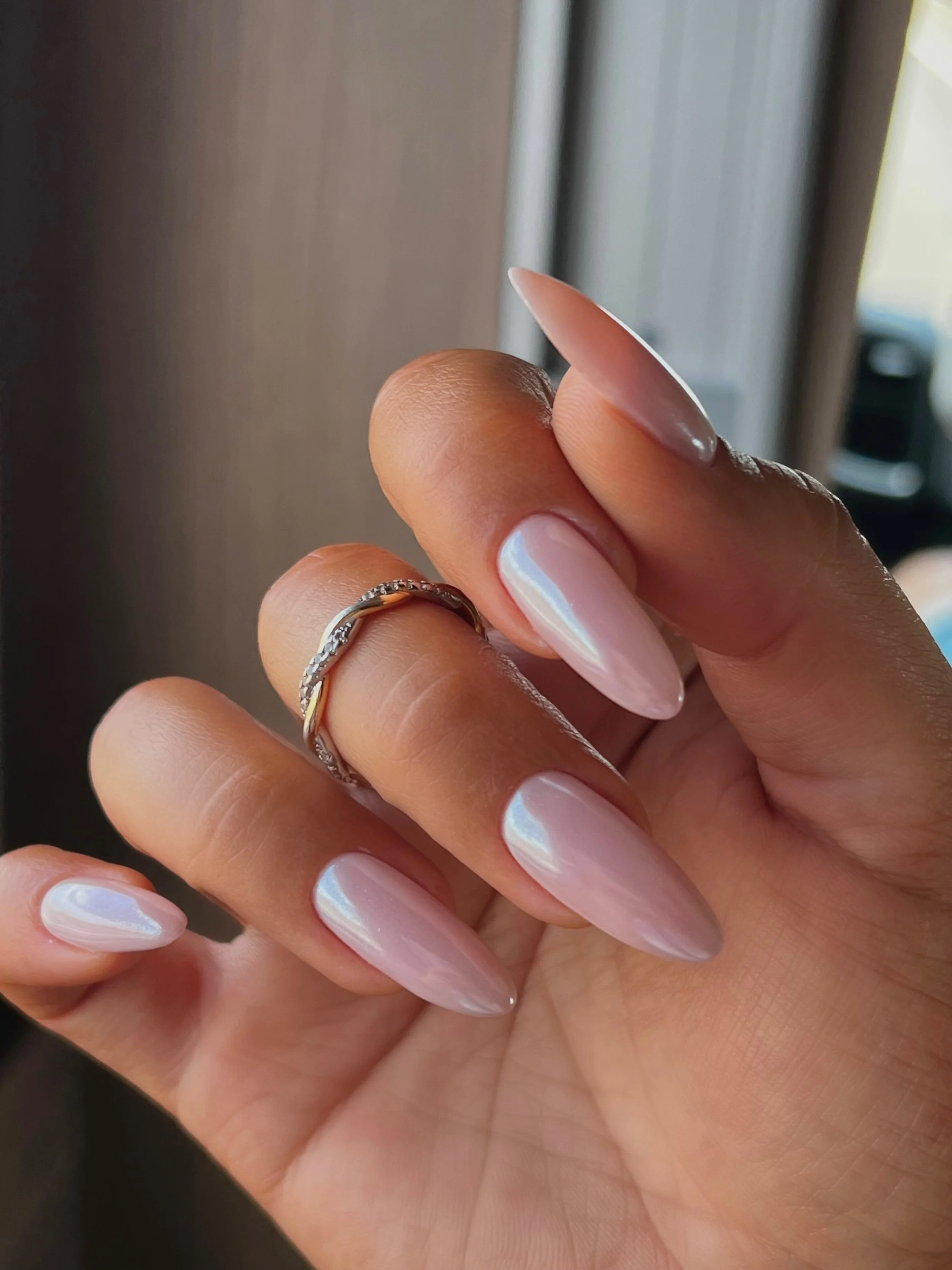 Close-up of a hand with long, almond-shaped nails painted in a pearly iridescent polish and wearing a twisted silver ring.
