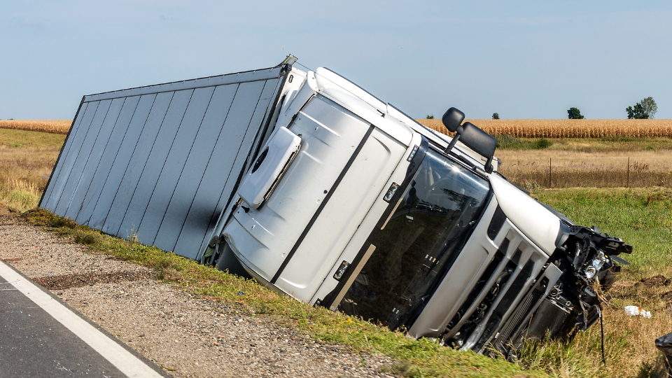 A semi-truck overturned on the side of a rural highway, with fields and a blue sky in the background.