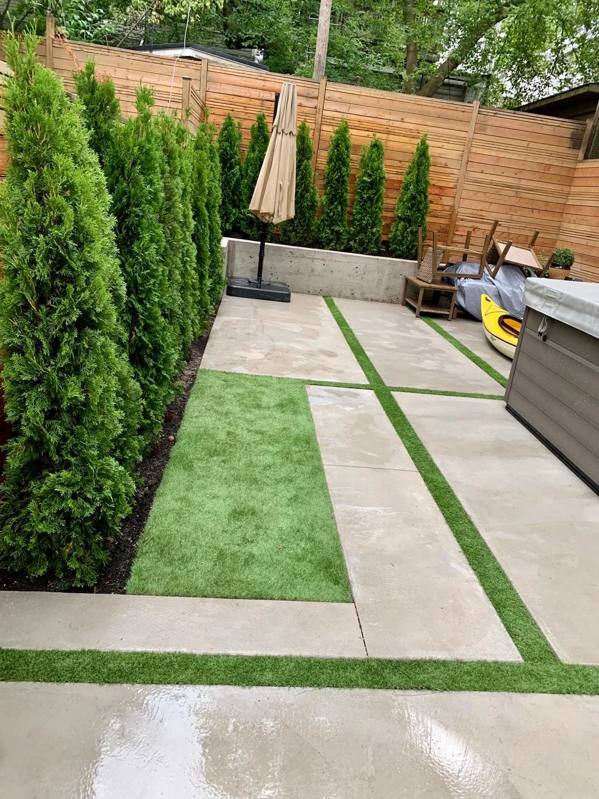 A backyard patio area with concrete slabs and artificial grass strips, bordered by a row of tall, dense green shrubs, a wooden fence, and a small storage shed.