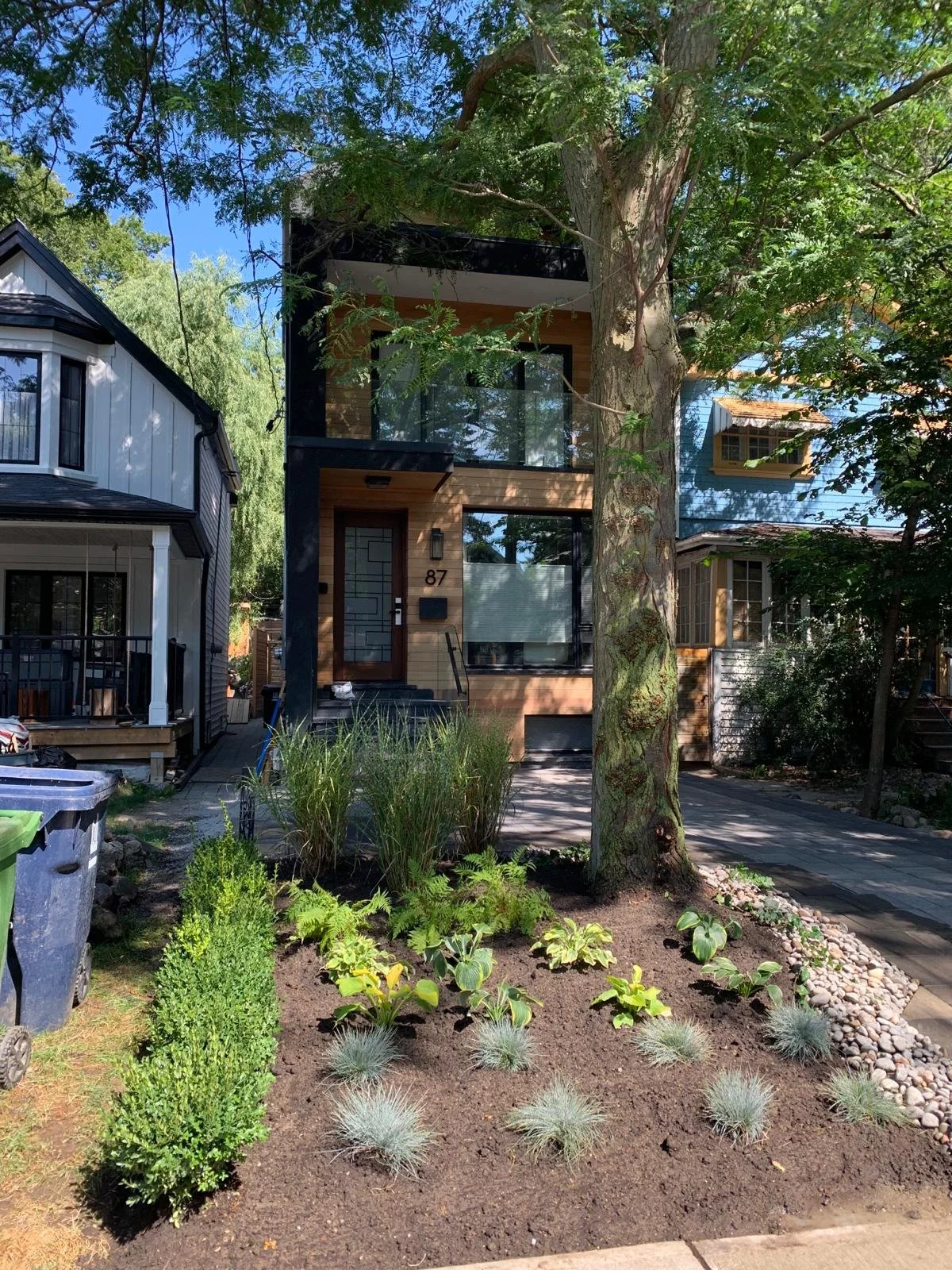 Front view of a modern two-story house with a glass balcony, surrounded by a landscaped yard with plants, bushes, and a large tree, on a sunny day.