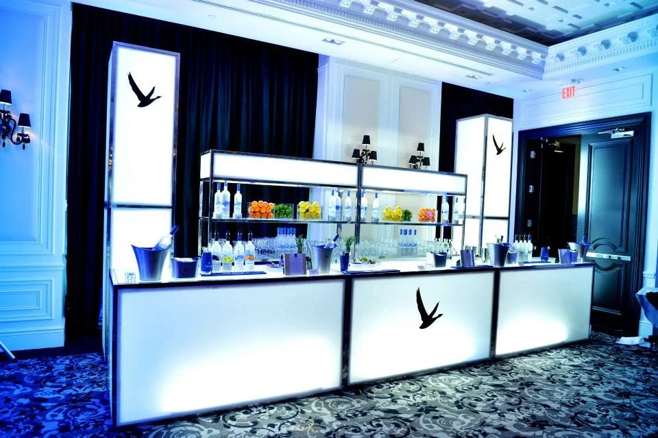 An illuminated bar setup with bottles, glasses, and decorative lemons, in a luxurious room with black curtains, wall sconces, and bird silhouettes on lit panels.