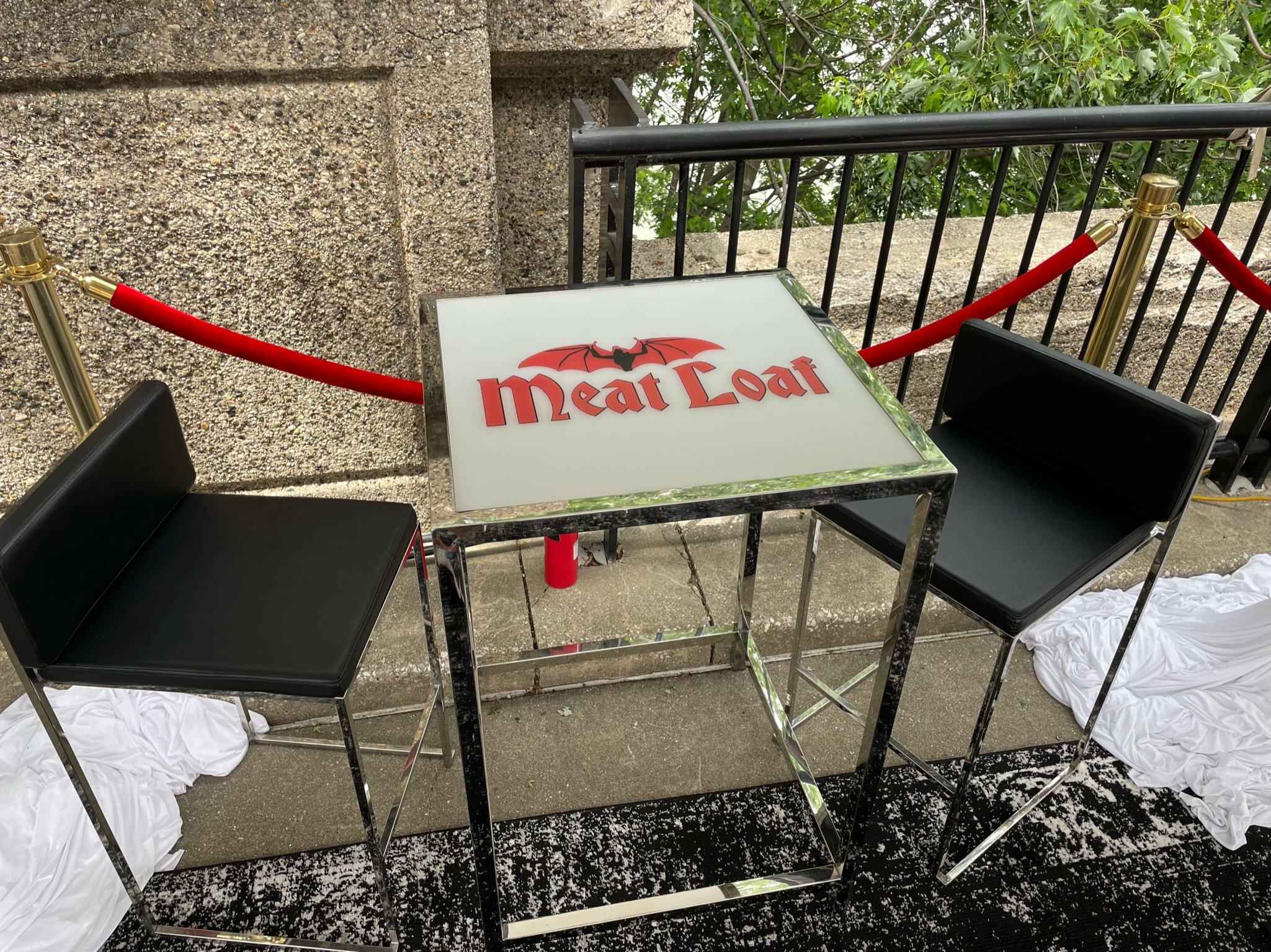 Table with a sign that reads 'Meat Load' featuring a bat with red wings and black body, surrounded by black chairs, red velvet chains as a barrier, and white fabric on the ground, with a stone wall and leafy trees in the background.
