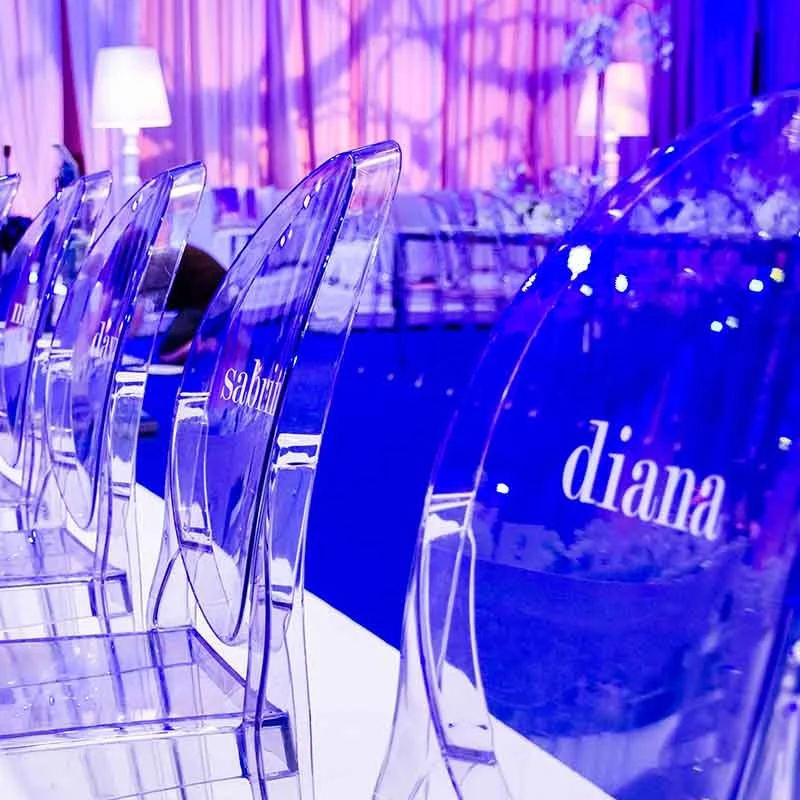A row of clear acrylic chairs with name tags in front of a decorated table, illuminated by purple and pink lighting, at an event or celebration.