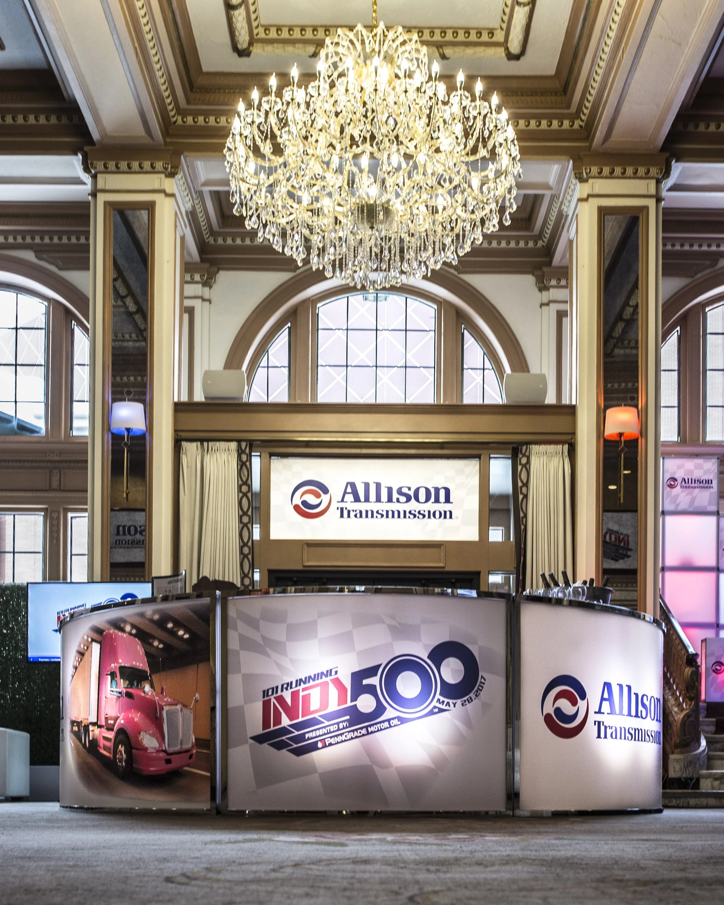 Indoor event space with a large chandelier hanging from ornate ceiling, banners displaying 'Allison Transmission' and an advertisement for an event with a pink truck and racing graphics.