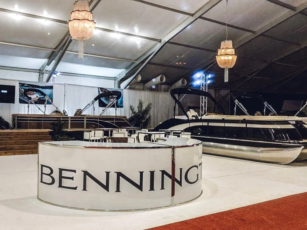 Luxury boats displayed in an indoor showroom with a white and black theme, white chairs, chandeliers, and a large sign with 'Benning' on it.
