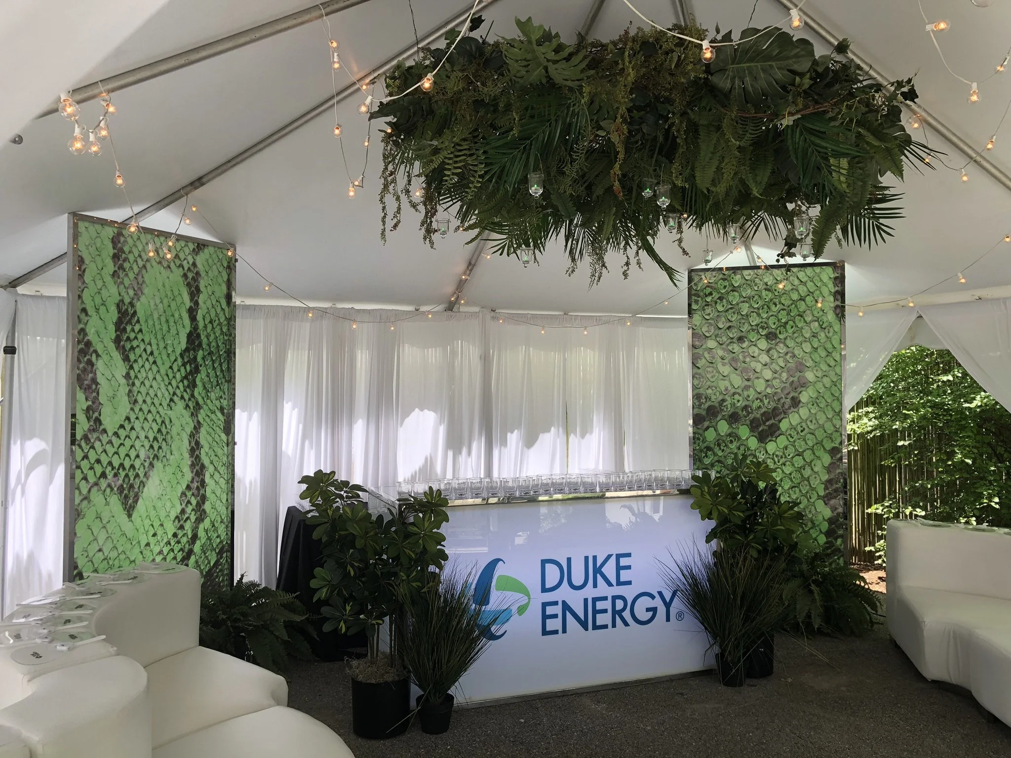 Inside a tent decorated with string lights, plants, and green patterned screens, featuring a sign with 'DUKE ENERGY' at an event.