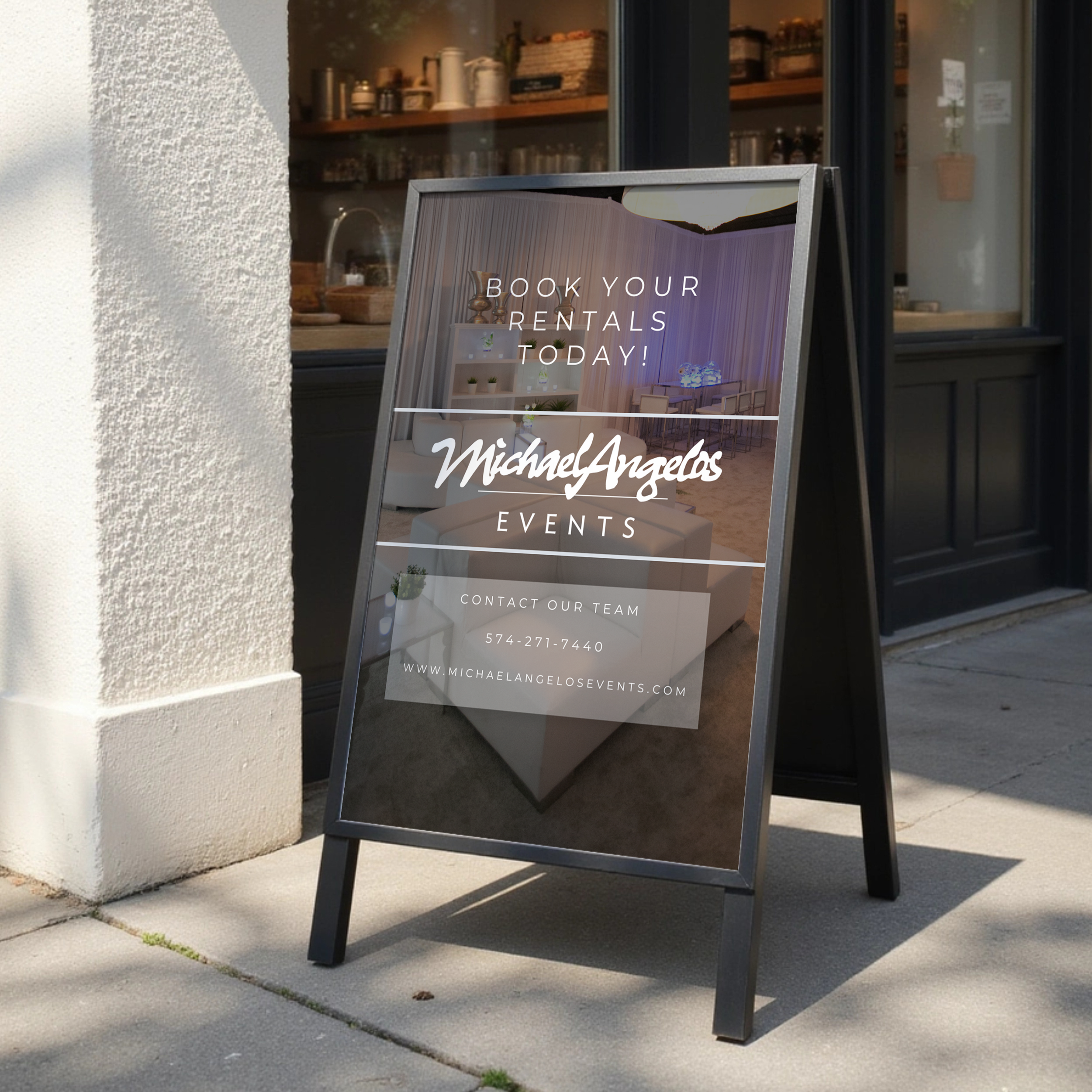 A sidewalk sign with a promotion for booking rentals by Michael Angelos Events, featuring interior decor images, placed outside a building with interior shelves visible through a window.