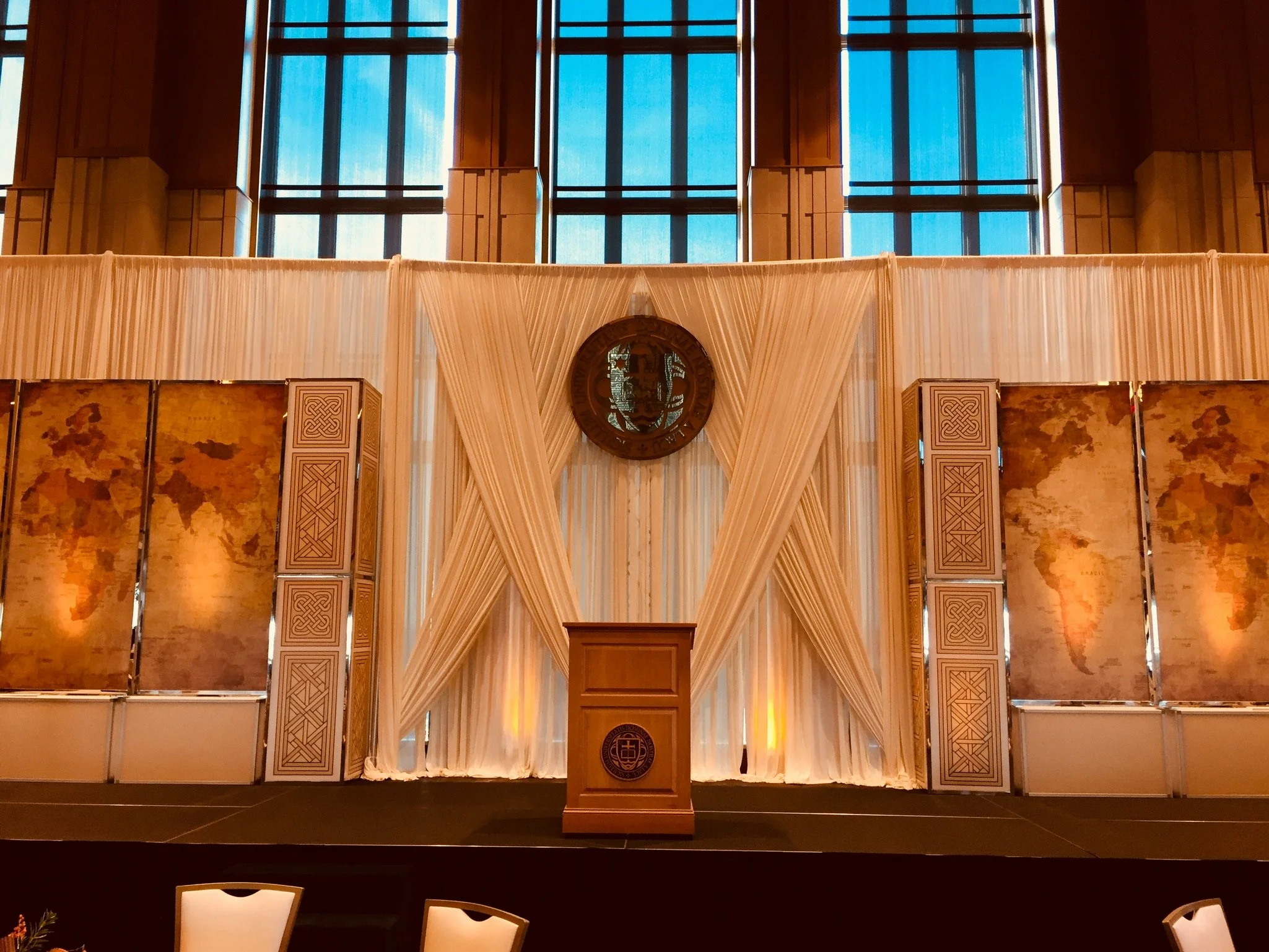 A large indoor stage with a wooden podium, decorated with a white curtain backdrop, gold accents, and a seal on the front, set up in a formal setting with tall windows and a high ceiling.
