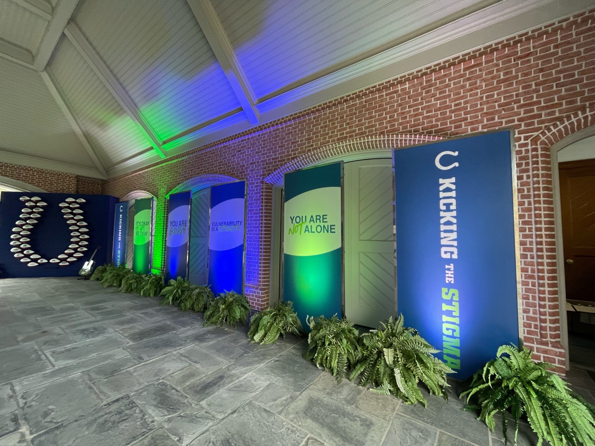 Indoor space with exposed brick walls and gray tile flooring. A row of vertical banners with inspirational messaging stands in front of the wall, adorned with green plants along the base. The ceiling is decorated with colored lighting.