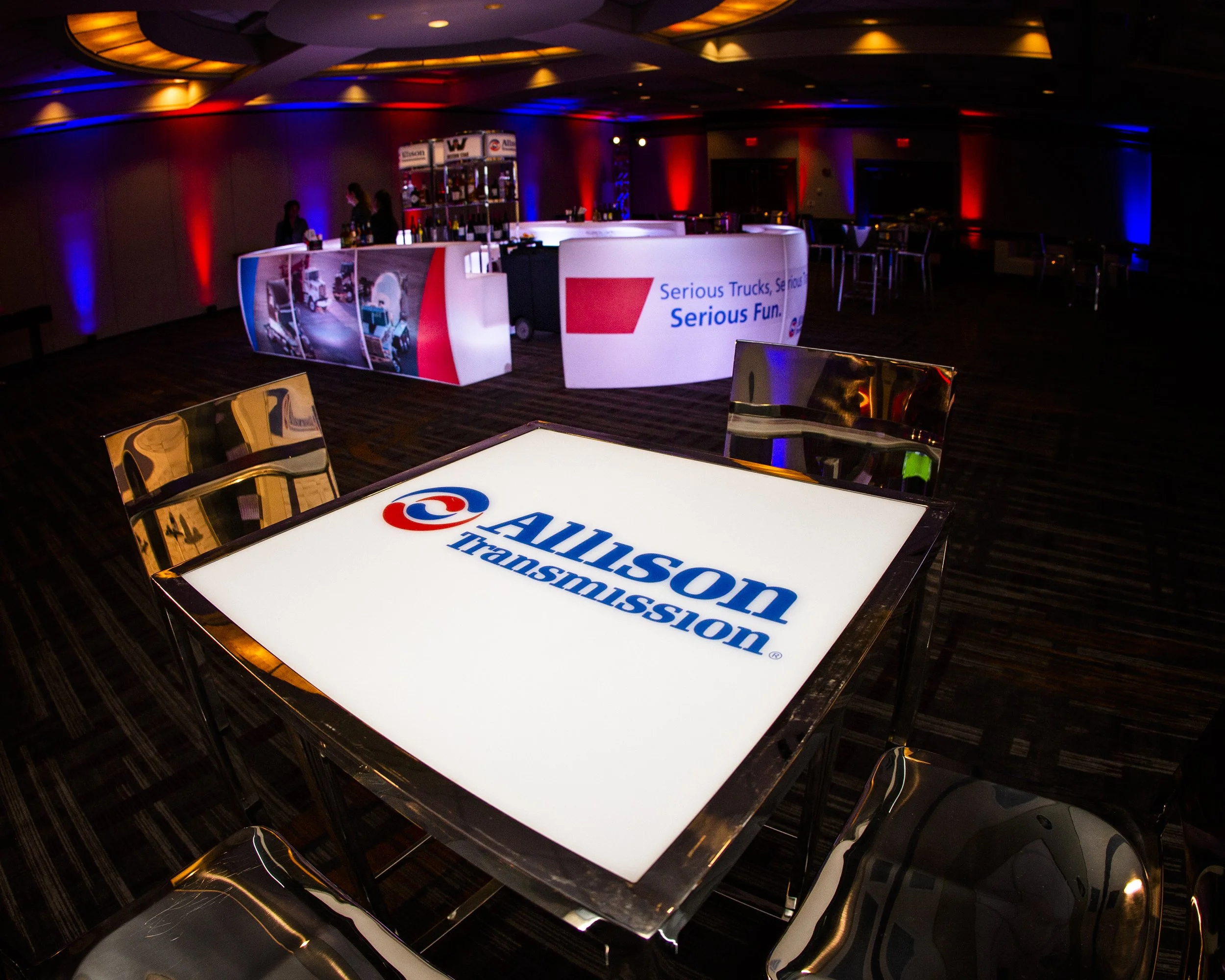 A dimly lit conference room or event space with tables featuring illuminated surfaces displaying the "ALLISON TRANSMISSION" logo. In the background, there's a booth with a sign that says "Serious Trucks, Serious Fun" and some people standing near it. The room is decorated with red and blue accent lighting on the walls.