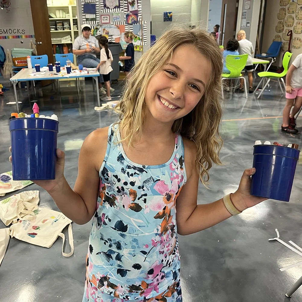 A young camper smiling and holding two containers of markers in a colorful classroom