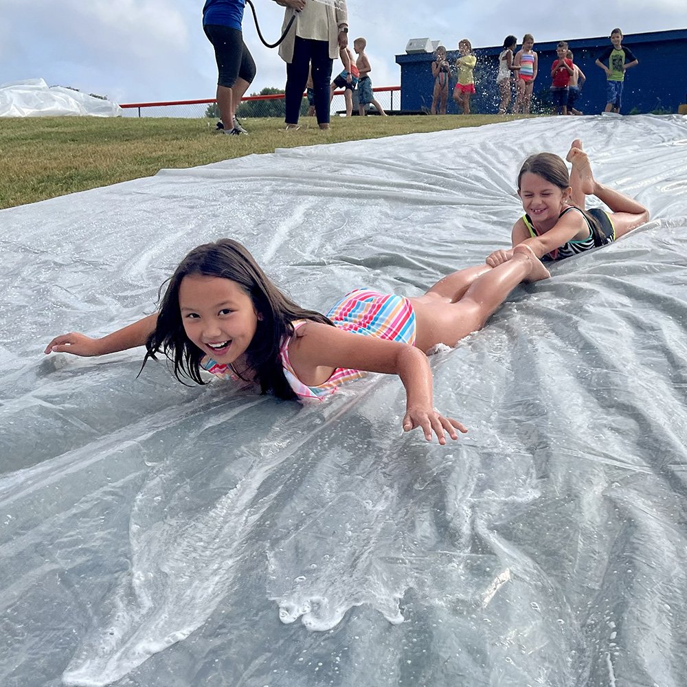 Two young campers smiling as they slide down a plastic water slide outdoors