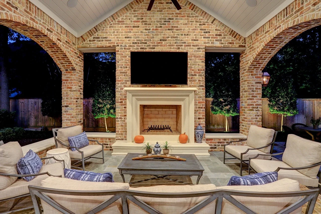 Seamless outdoor living space with a custom brick patio cover, arched architectural details, pavers, and a cast stone fireplace, designed to look original to the home and placed alongside a pool and spa with fountains.