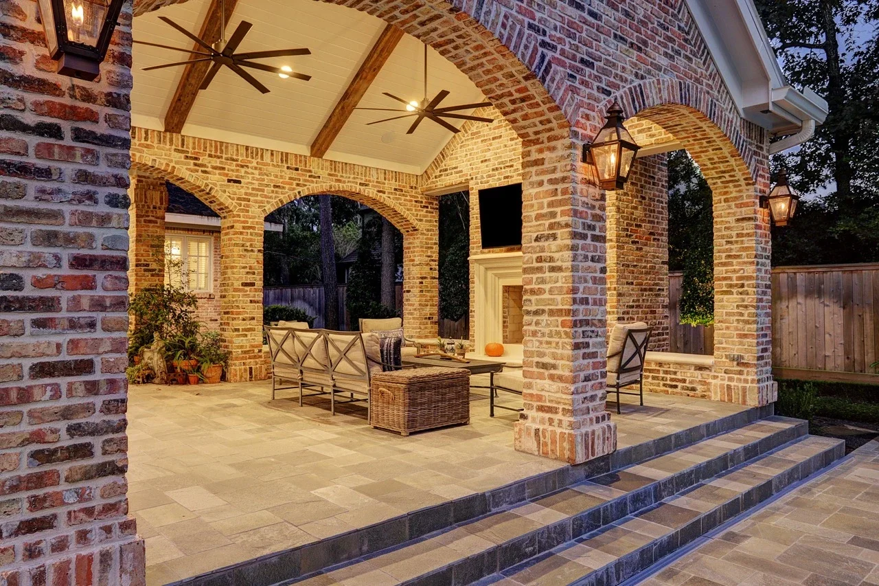Seamless outdoor living space with a custom brick patio cover, arched architectural details, pavers, and a cast stone fireplace, designed to look original to the home and placed alongside a pool and spa with fountains.