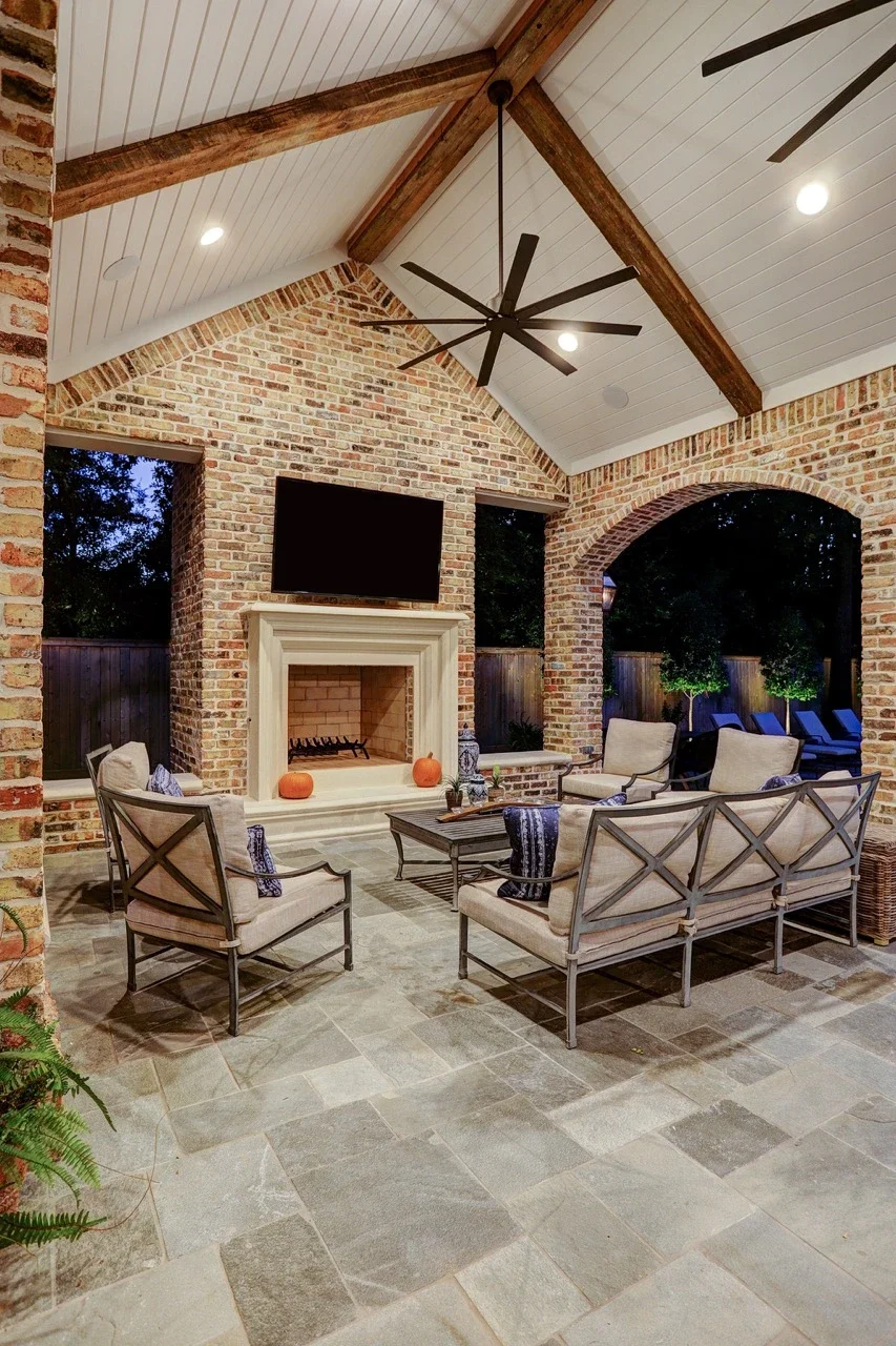 Seamless outdoor living space with a custom brick patio cover, arched architectural details, pavers, and a cast stone fireplace, designed to look original to the home and placed alongside a pool and spa with fountains.