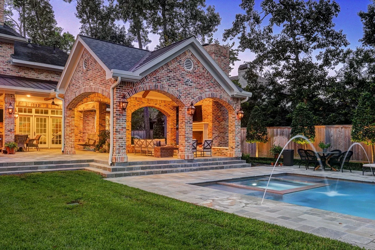 Seamless outdoor living space with a custom brick patio cover, arched architectural details, pavers, and a cast stone fireplace, designed to look original to the home and placed alongside a pool and spa with fountains.