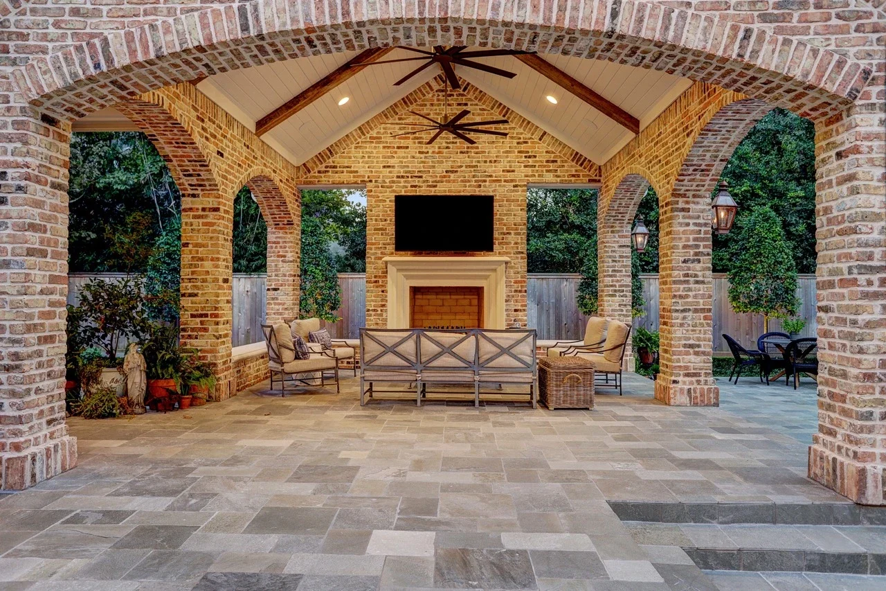 Seamless outdoor living space with a custom brick patio cover, arched architectural details, pavers, and a cast stone fireplace, designed to look original to the home and placed alongside a pool and spa with fountains.