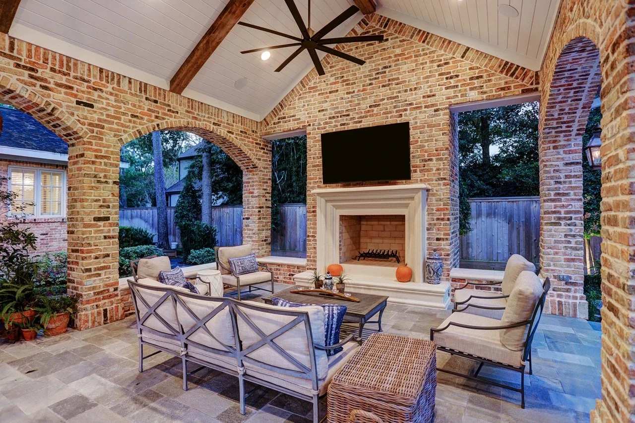 Seamless outdoor living space with a custom brick patio cover, arched architectural details, pavers, and a cast stone fireplace, designed to look original to the home and placed alongside a pool and spa with fountains.
