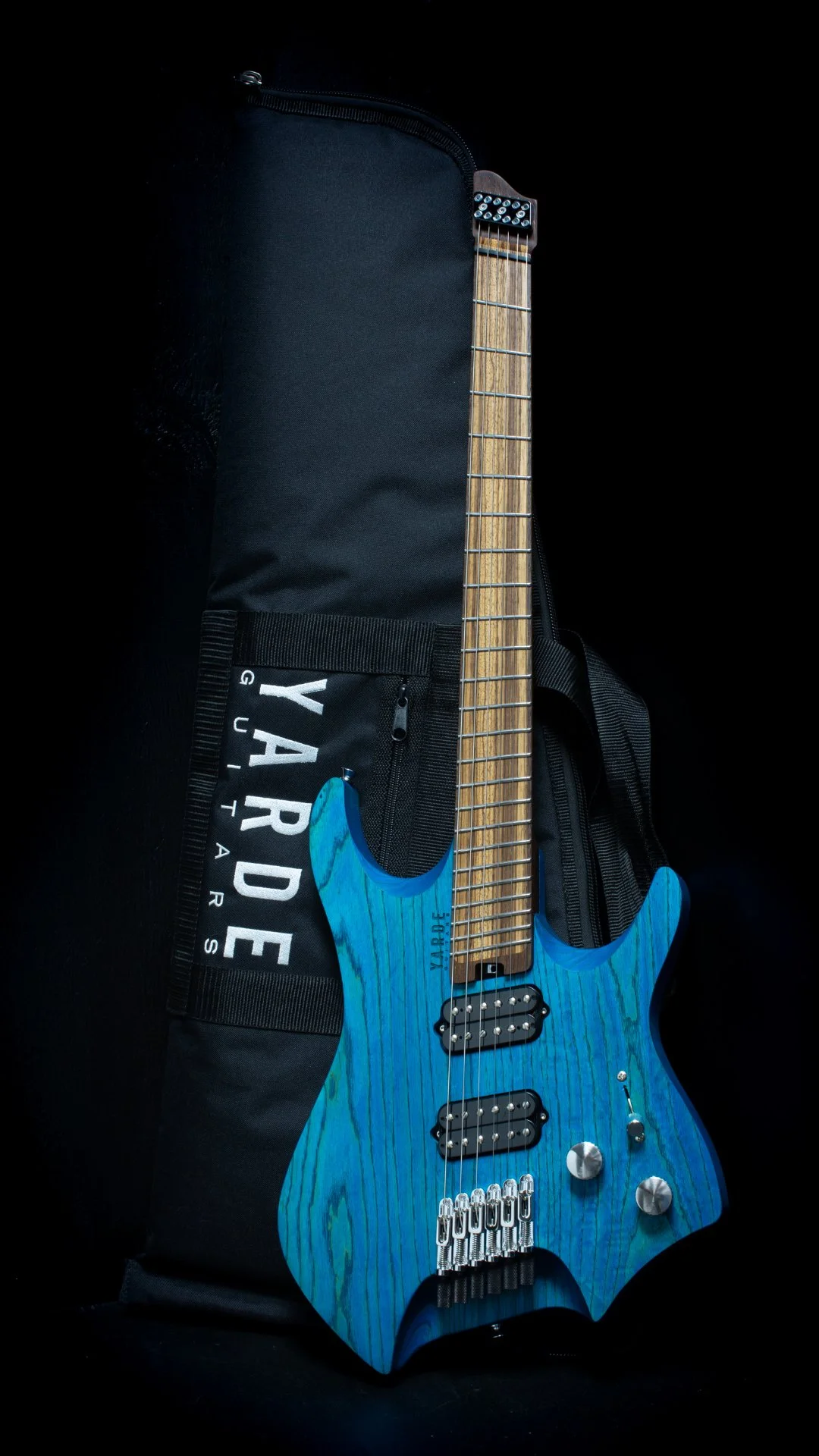 A blue stained headless - multiscale electric guitar with a two angled humbuckers and chrome hardware leaning against a 'Yarde Guitars' branded black gig-bag, against a black background.