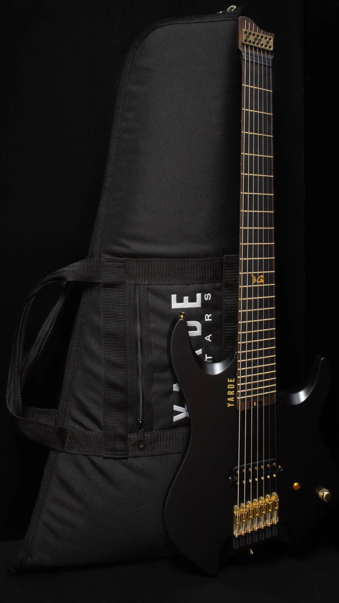 A black headless - multiscale electric guitar with a single humbucker and gold hardware leaning against a 'Yarde Guitars' branded black gig-bag, against a black background.