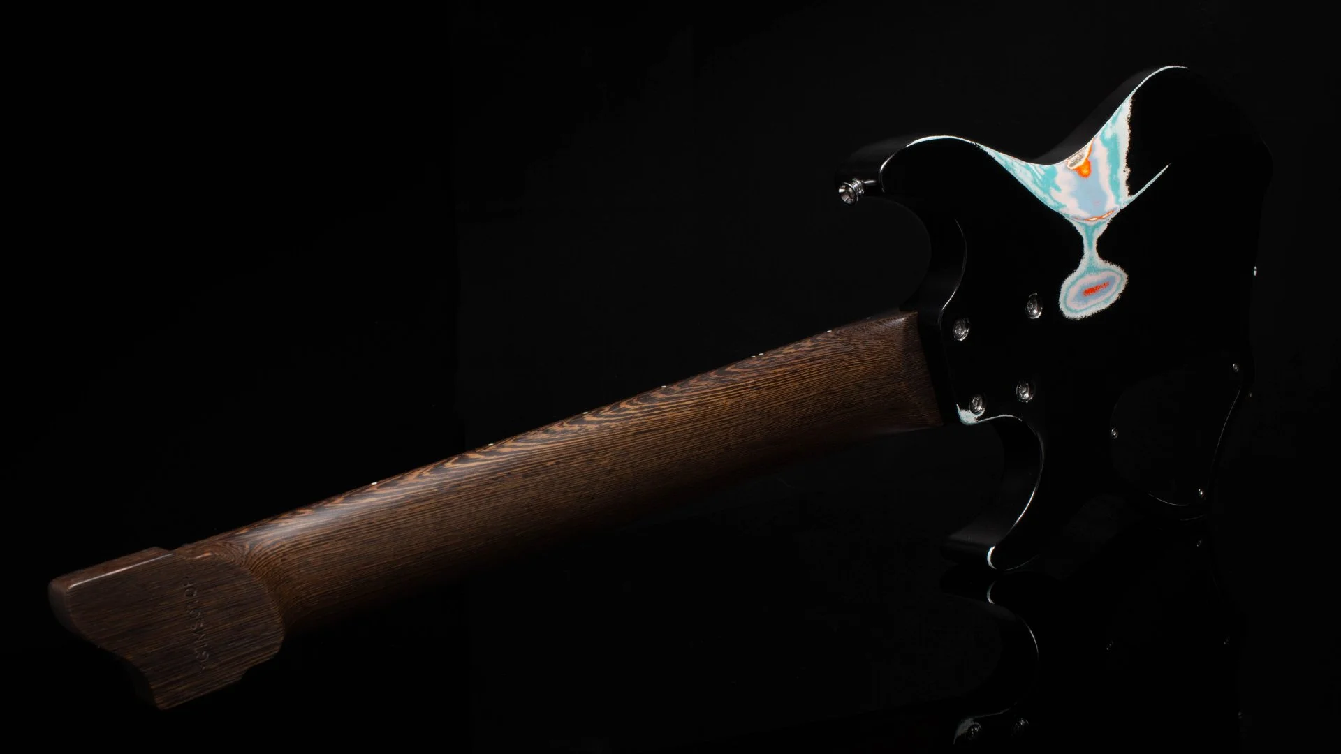 Back view of an 8 string headless - multisclae electric guitar with black paint and multi coloured relic patterns
