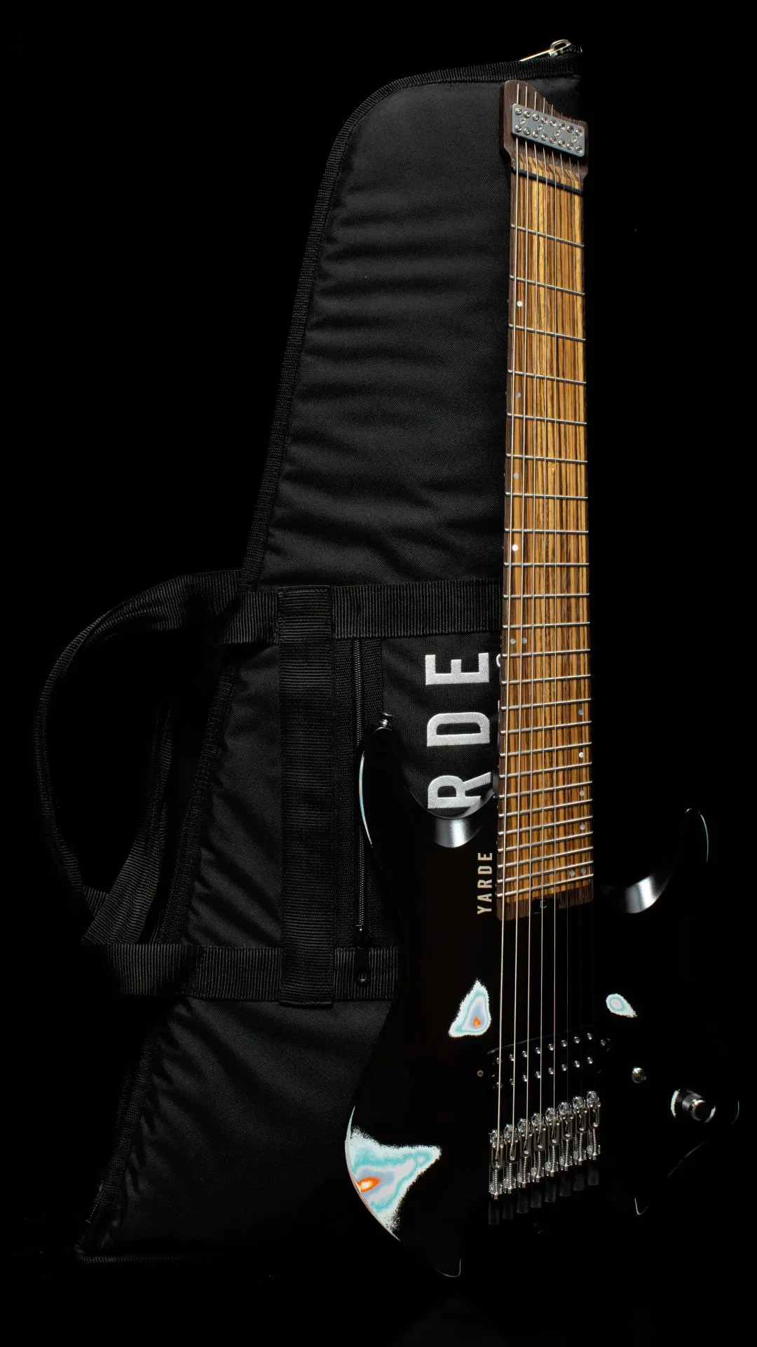A 8 string headless - multiscale electric guitar leaning on a 'Yarde Guitars' branded gig bag against a black back ground