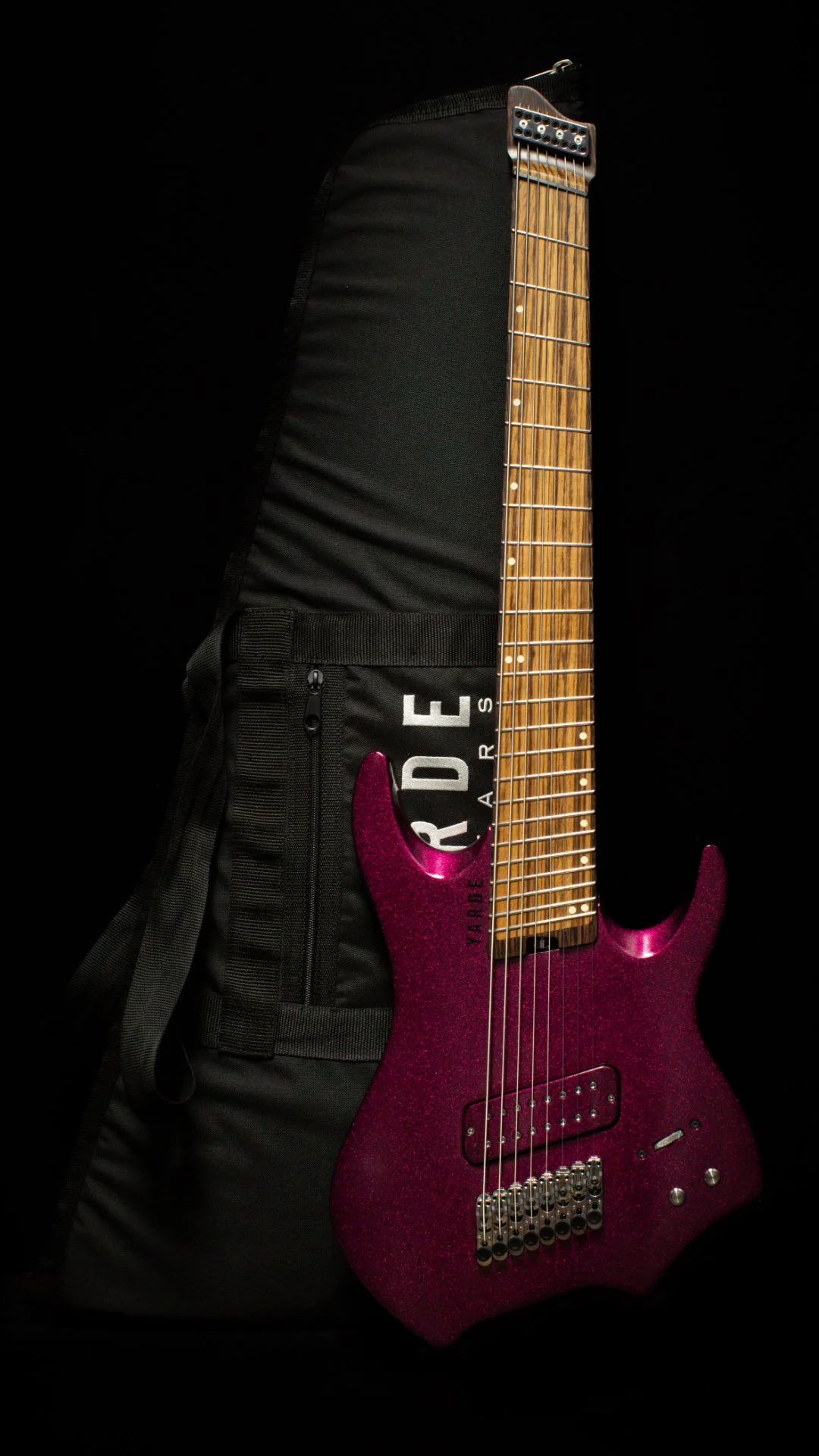 A 8 string headless guitar with a pink sparkle finish leaning on a 'Yarde Guitars' against a black background