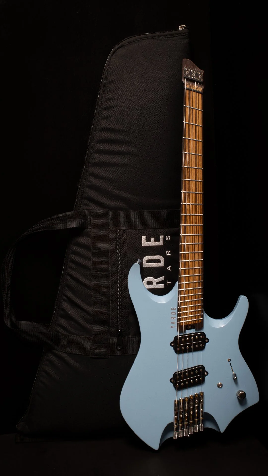 A light blue headless - multiscale electric guitar with custom humbucker pickups and chrome hardware leaning against a 'Yarde Guitars' branded black gig-bag, against a black background.