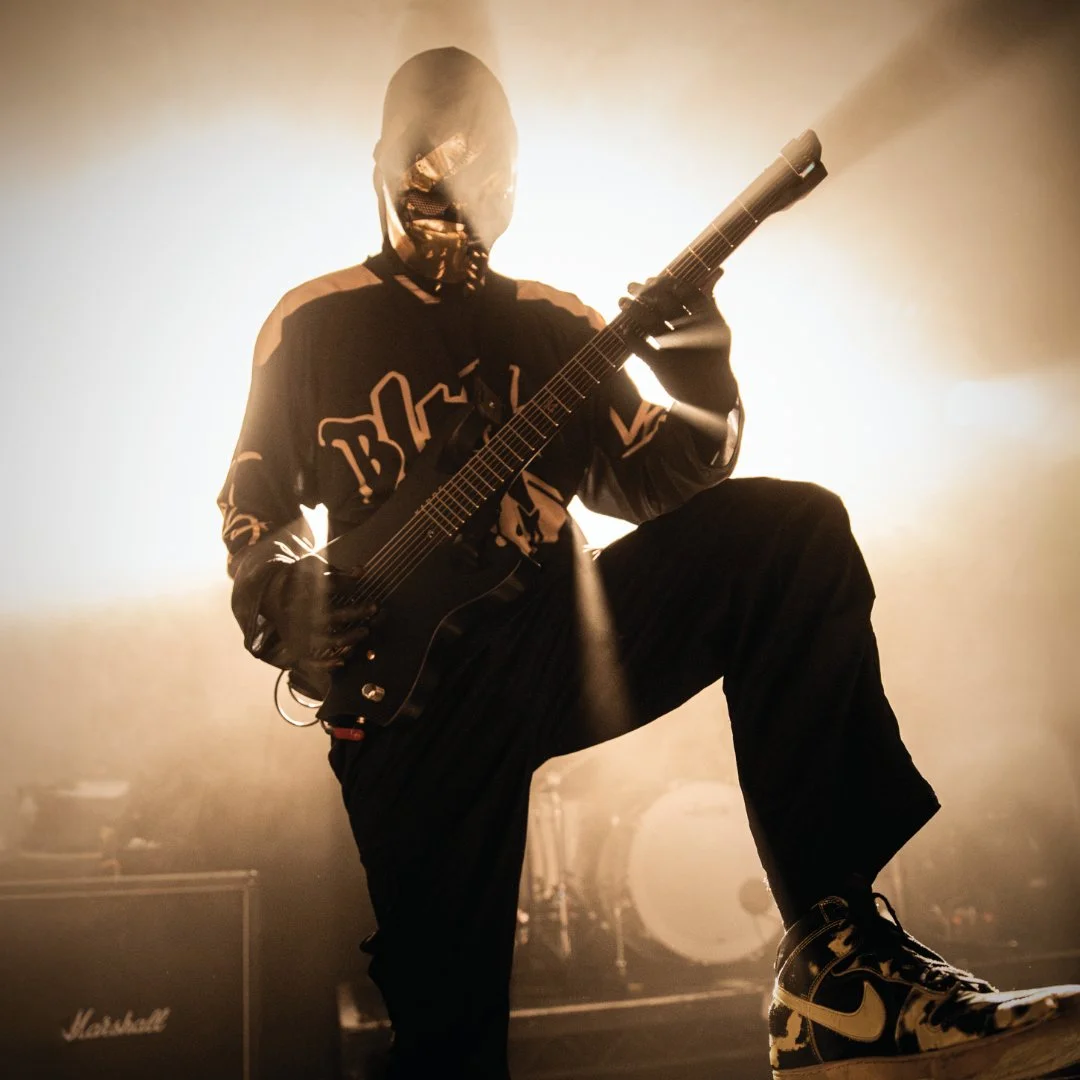 Nu-Metal Guitarist 'JUDGE' of BLACKGOLD wearing a mask playing a custom electric guitar on stage with a backlit haze, Marshall amplifier in the background.