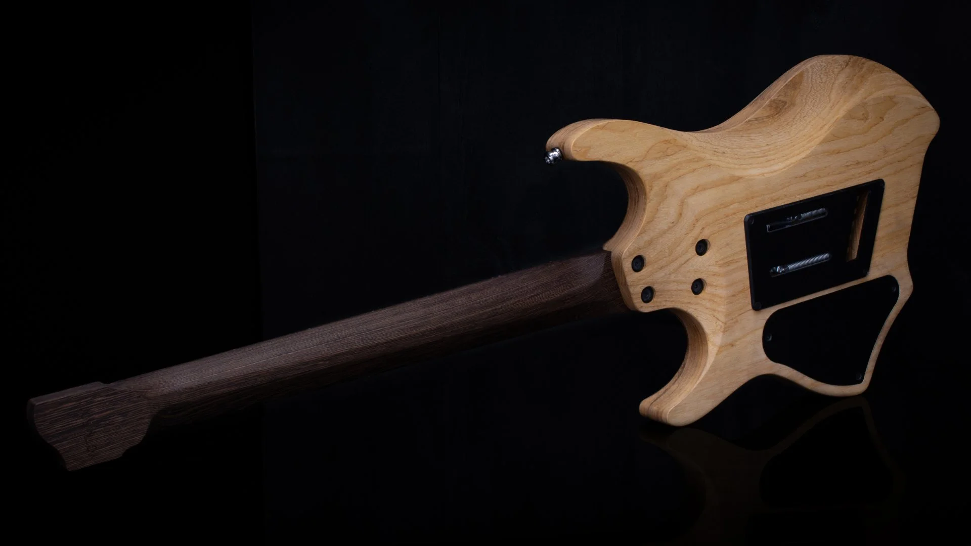 Close-up of the back of a headless - multiscale electric guitar with a swamp ash body and wenge neck and tremolo bridge against a black background - Landscape back view