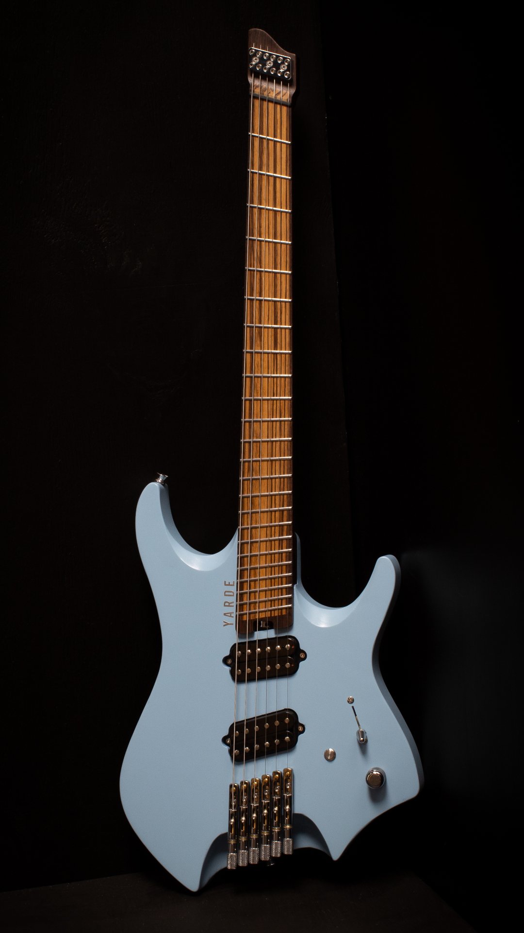 A headless - multiscale electric guitar with a light blue nitrocellulose finish, two custom humbucker pickups, a fixed bridge, wenge neck and zebrano fretboard, against a black background - Portrait view