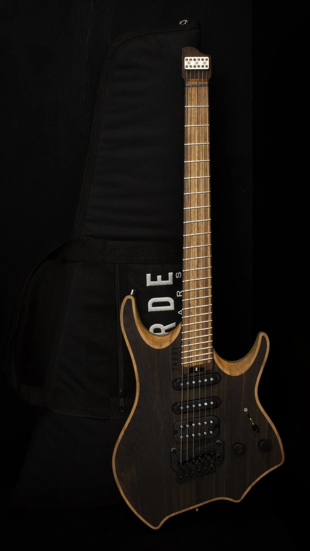A natural finished headless - multiscale electric guitar with a HSS pickups and black hardware leaning against a 'Yarde Guitars' branded black gig-bag, against a black background.