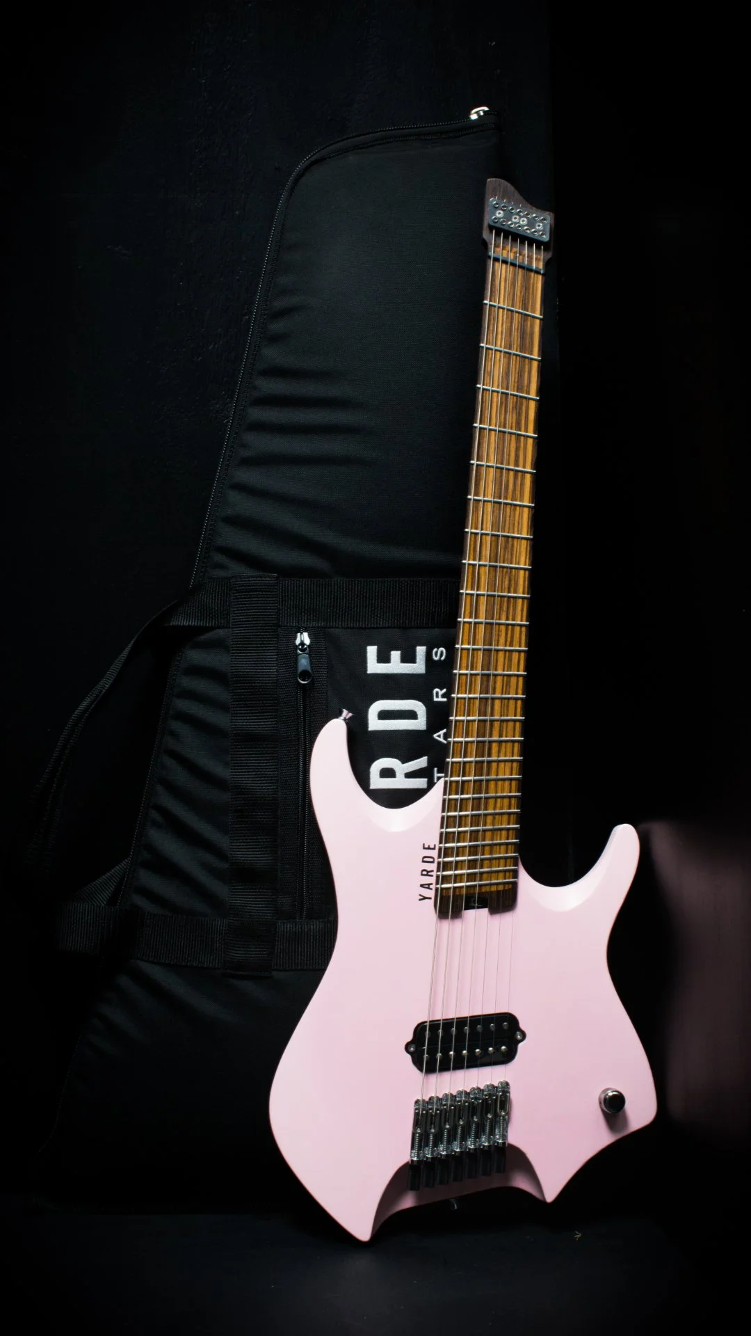 A pink headless - multiscale electric guitar leaning on a 'Yarde Guitars' branded gig bag against a black background