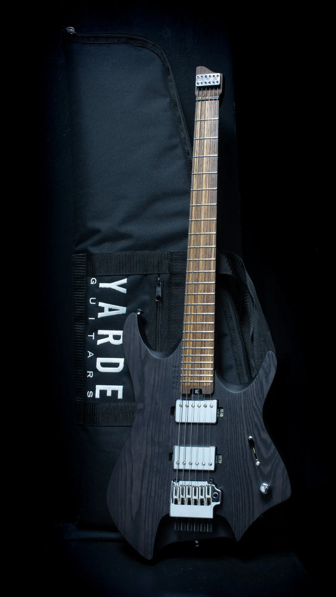 A black stained headless electric guitar leaning on a 'Yarde Guitars' branded gig bag against a black background