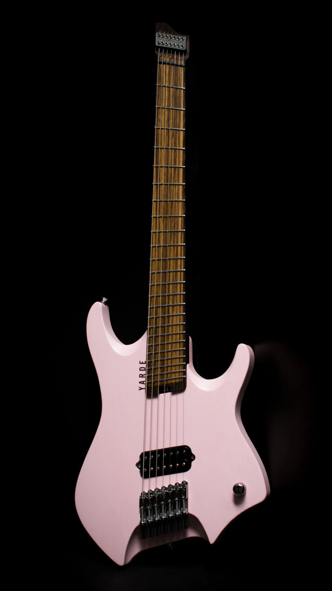 A pink painted 7 string headless - multiscale electric guitar, one humbucker pickup, chrome hardware, Wenge neck and inlaid zebrano fretboard, against a black background - Portrait view