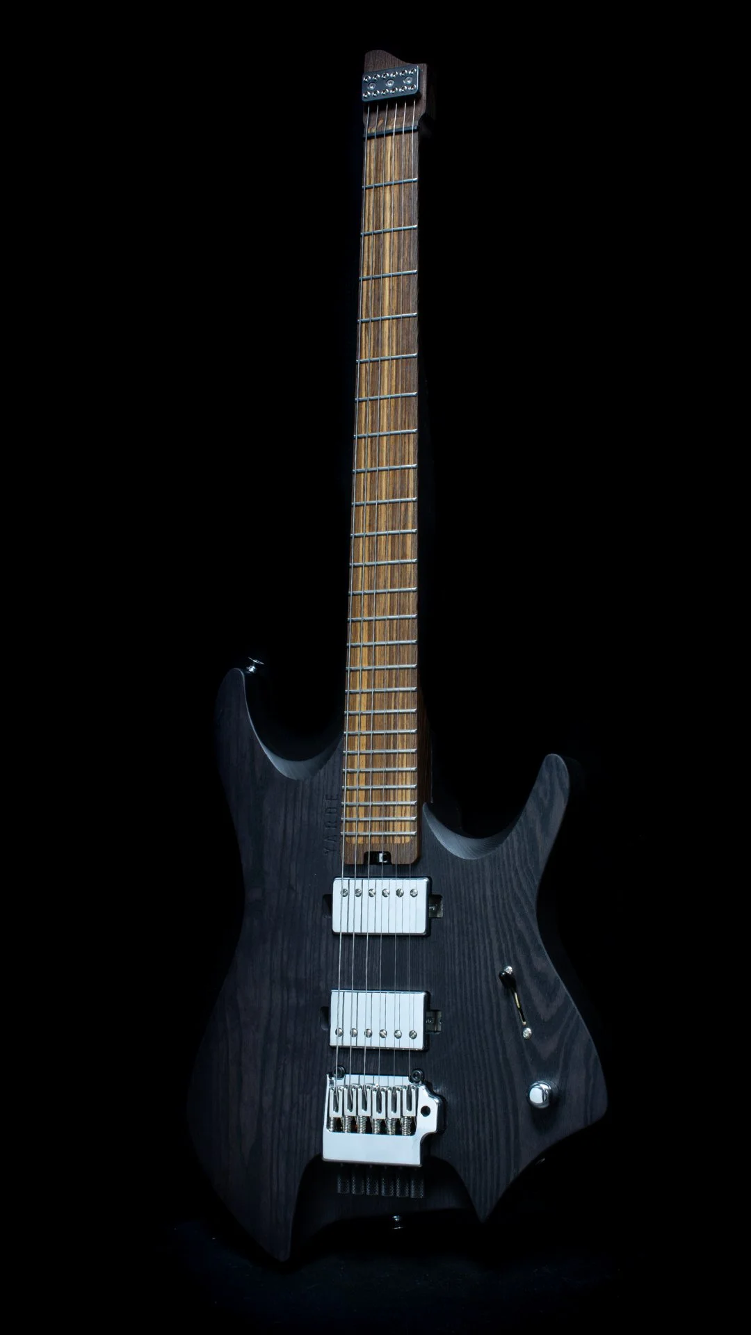 A headless guitar with a black stained wood finish, two humbucker pickups, a chrome tremolo bridge, Wenge neck and inlaid zebrano fretboard, against a black background - Portrait view