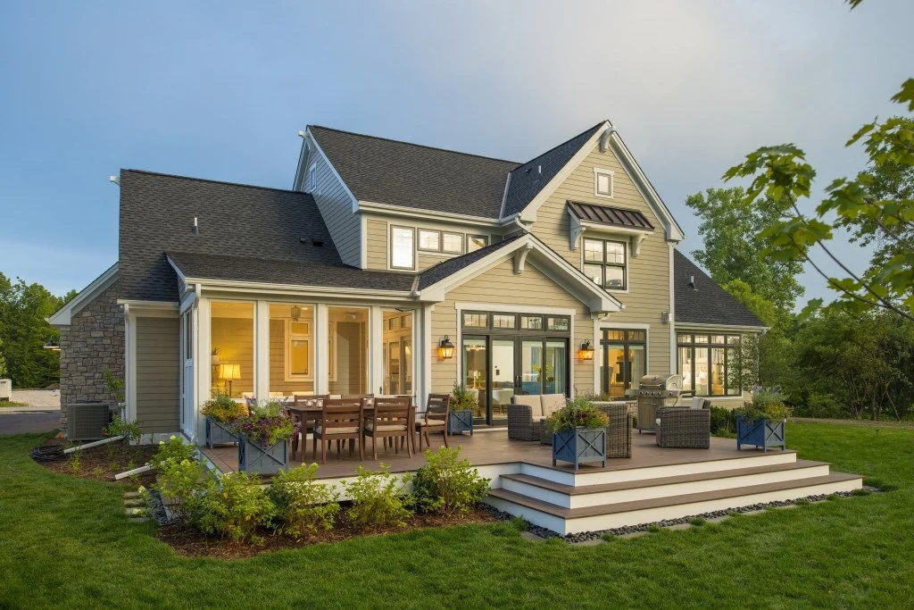 A large two-story house with a deck and outdoor furniture, surrounded by a green lawn and trees.
