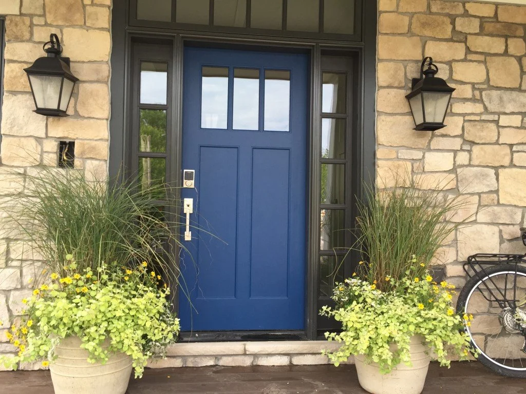 The image shows a front door with a deep blue color, set in a stone wall, flanked by two black outdoor lantern lights. Two large planters with green grasses and yellow flowers are placed on either side of the door. A bicycle is partially visible on t