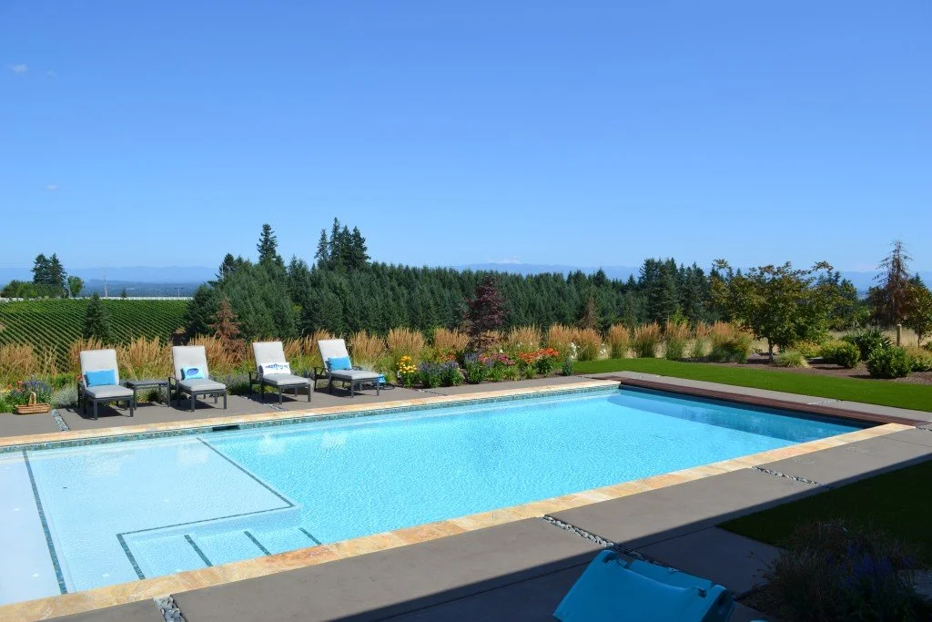 A swimming pool with lounge chairs on the pool deck, surrounded by a well-maintained garden and trees, with hills and mountains in the background under a clear blue sky.