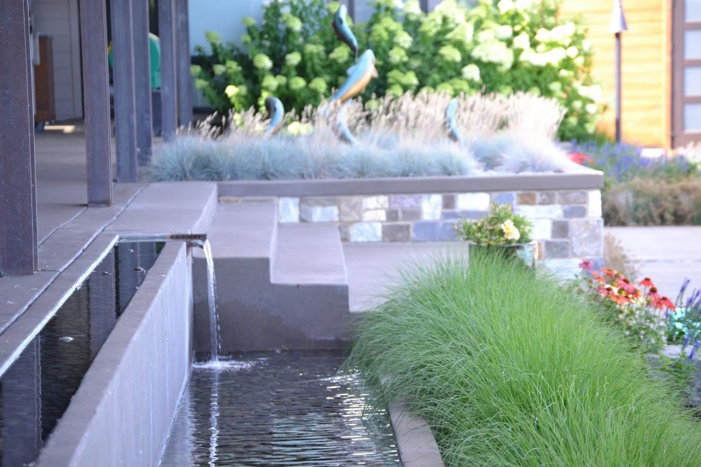 A backyard scene with a water feature and decorative garden plants, including a fountain, lush grass, flowers, and a small potted plant.