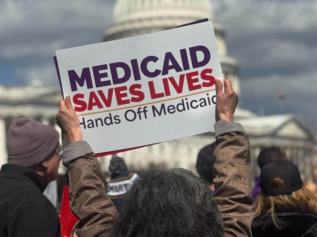 Person holding a sign that reads "Medicaid Saves Lives, Hands Off Medicaid" at a protest or rally near a government building.