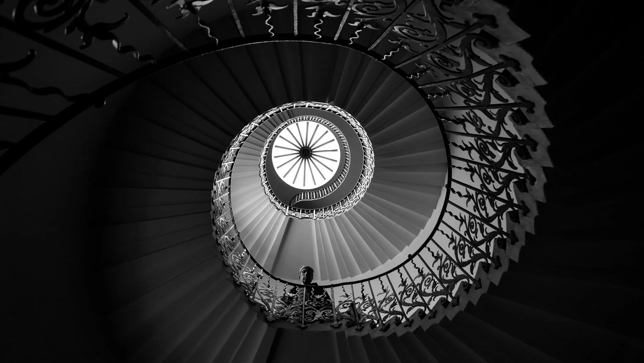 Black and white photo of a spiral staircase viewed from below, showing intricate railing and steps leading upward, with light coming from the top.